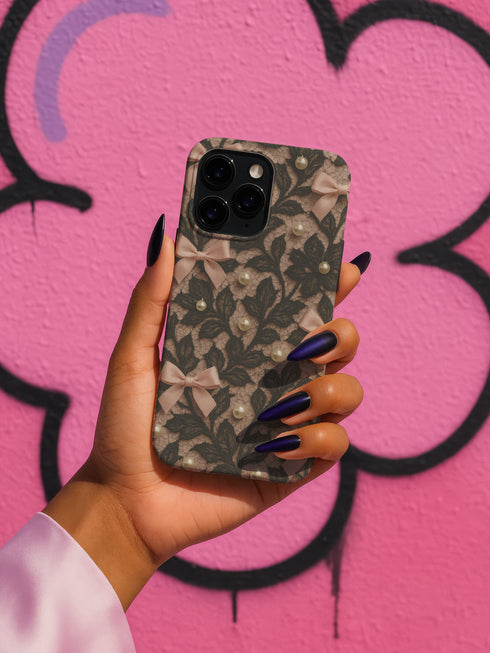 Phone case with black lace floral design, soft pink satin bows, and small pearl accents on a pale nude background; fits iPhone, Samsung, and Pixel models